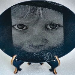 Oval black plaque on stand featuring an etched grayscale close-up of a child's face with large eyes.