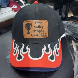 A black and red cap with flame design and a patch that reads "Pine Valley Trophy Case, LLC" with a trophy icon.