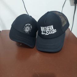 Two black trucker hats on a brown surface; one reads "MUSTAPHA 2024" and the other "NORTHERN Elite.